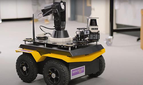 A robot car inside a lab with four thick wheels a yellow chassis and equipment on top