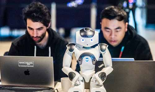 Two men looking at laptop with a humanoid robot in-between them looking directly at the camera