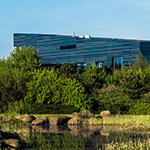 A modern building, the Dalton Cumbrian Facility, surrounded by greenery, with a pond in the foreground reflecting the sky.