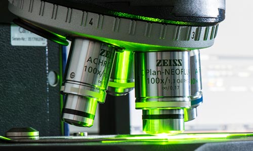 A close-up of a microscope with green illumination, analysing a sample with a computer screen displaying magnified results in the background.