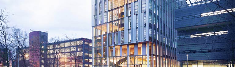 A modern glass-fronted building, the Royce Hub, surrounded by trees and people in an urban setting.